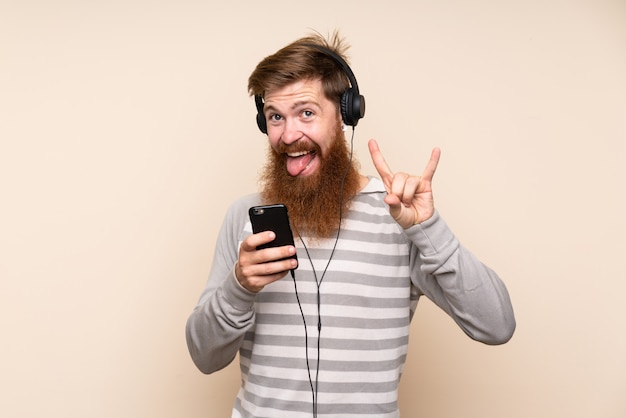 Hombre pelirrojo con barba larga sobre fondo aislado usando el móvil con auriculares y cantando