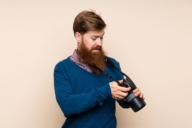 Hombre pelirrojo con barba larga sobre pared aislada con una cámara profesional