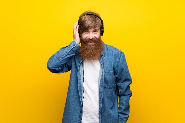 Hombre pelirrojo con barba larga sobre pared amarilla aislada escuchando música con auriculares