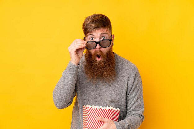 Hombre pelirrojo con barba larga sobre pared amarilla aislada sorprendido con gafas 3d y sosteniendo un gran cubo de palomitas de maíz