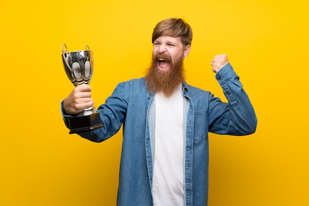 Foto hombre pelirrojo con barba larga sobre pared amarilla aislada sosteniendo un trofeo