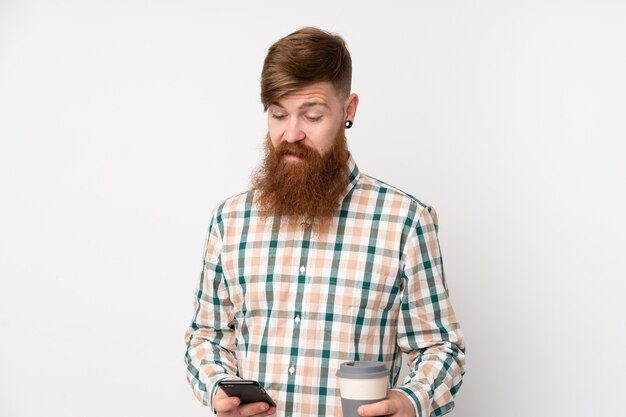 Hombre pelirrojo con barba larga sobre pared blanca aislada con café para llevar y un móvil
