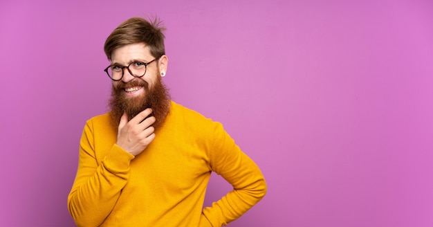 Hombre pelirrojo con barba larga sobre pared púrpura con gafas y sonriente