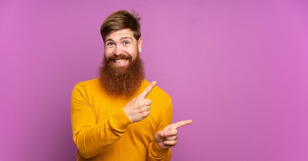 Hombre pelirrojo con barba larga sobre púrpura aislado asustado y apuntando hacia un lado