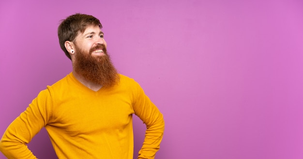 Hombre pelirrojo con barba larga sobre púrpura aislado posando con los brazos en la cadera y sonriendo