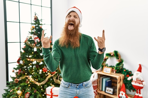 Hombre pelirrojo con barba larga con sombrero de navidad junto al árbol de navidad gritando con expresión loca haciendo símbolo de rock con las manos arriba. estrella de la música concepto de música pesada.