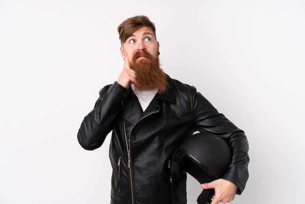 Hombre pelirrojo con barba larga sosteniendo un casco de moto sobre pared blanca pensando una idea