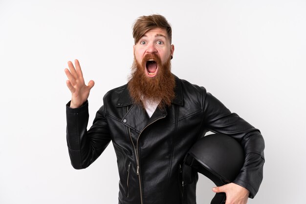 Hombre pelirrojo con barba larga sosteniendo un casco de motocicleta sobre blanco aislado con expresión facial sorpresa