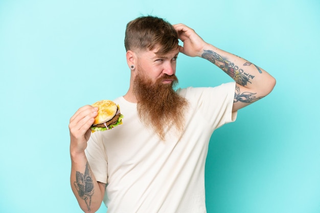 Hombre pelirrojo con barba larga sosteniendo una hamburguesa aislada de fondo azul con dudas y con expresión facial confusa
