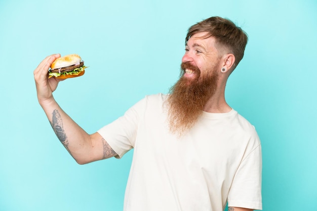 Hombre pelirrojo con barba larga sosteniendo una hamburguesa aislada de fondo azul con expresión feliz