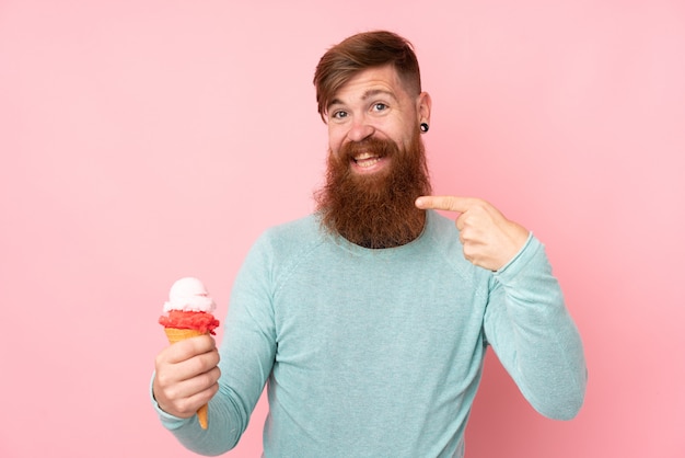 Hombre pelirrojo con barba larga sosteniendo un helado de cucurucho sobre pared rosa aislado y apuntando