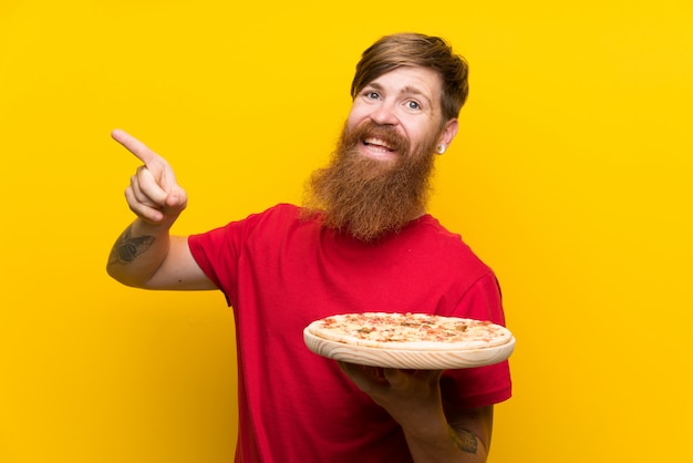 Hombre pelirrojo con barba larga sosteniendo una pizza sobre pared amarilla aislada sorprendido y apuntando con el dedo hacia un lado