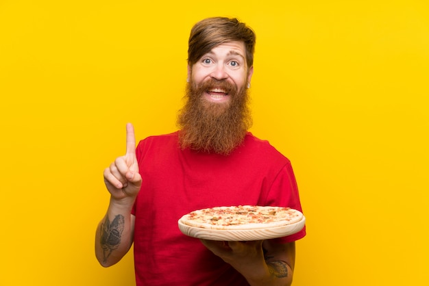 Hombre pelirrojo con barba larga sosteniendo una pizza sobre pared amarilla apuntando hacia una gran idea