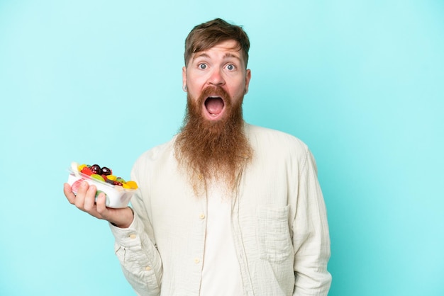 Hombre pelirrojo con barba larga sosteniendo un tazón de fruta aislado de fondo azul con expresión facial sorprendida y sorprendida