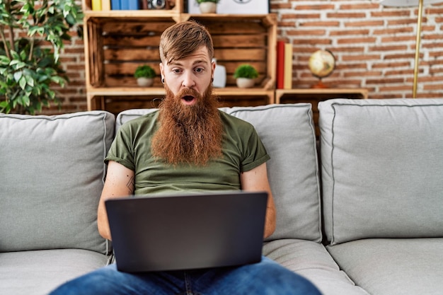 Hombre pelirrojo con barba larga usando una computadora portátil sentado en el sofá en la sala de estar asustado y asombrado con la boca abierta por sorpresa, cara de incredulidad