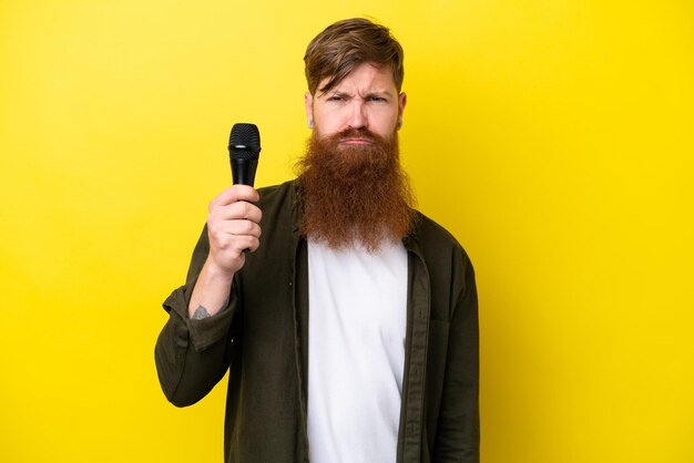 Hombre pelirrojo con barba recogiendo un micrófono aislado de fondo amarillo con expresión triste