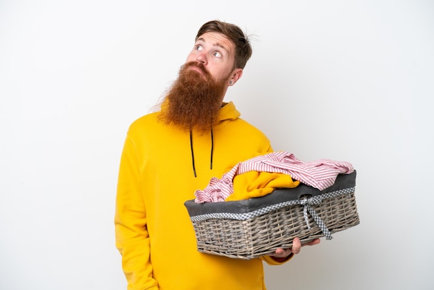 Hombre pelirrojo con barba sosteniendo una cesta de ropa aislada de fondo blanco y mirando hacia arriba