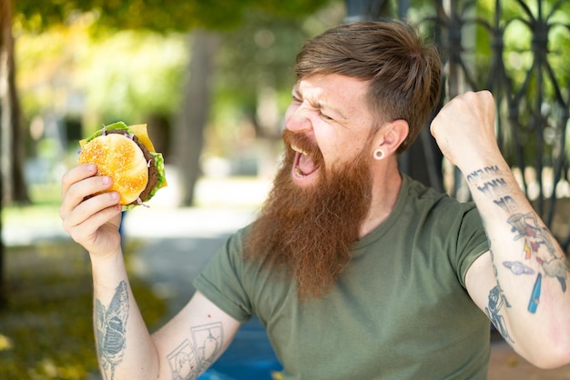 Hombre pelirrojo con barba sosteniendo una hamburguesa al aire libre celebrando una victoria