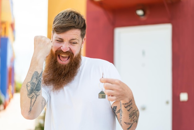 Hombre pelirrojo con barba sosteniendo llaves de casa al aire libre celebrando una victoria