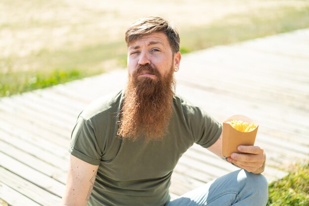 Hombre pelirrojo con barba sosteniendo papas fritas al aire libre haciendo gesto de dudas mientras levanta los hombros