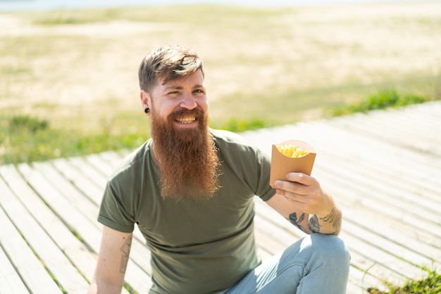 Hombre pelirrojo con barba sosteniendo papas fritas al aire libre sonriendo mucho