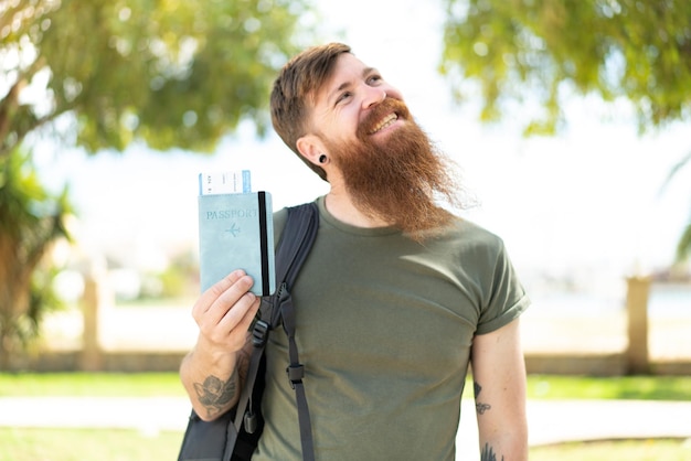 Hombre pelirrojo con barba sosteniendo un pasaporte al aire libre mirando hacia arriba mientras sonríe