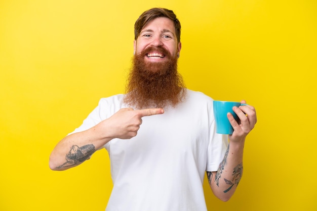 Hombre pelirrojo con barba sosteniendo una taza aislada de fondo amarillo y señalándola