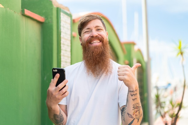 Hombre pelirrojo con barba usando teléfono móvil al aire libre apuntando hacia un lado para presentar un producto