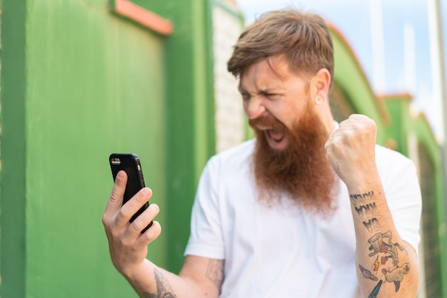 Hombre pelirrojo con barba usando teléfono móvil al aire libre celebrando una victoria