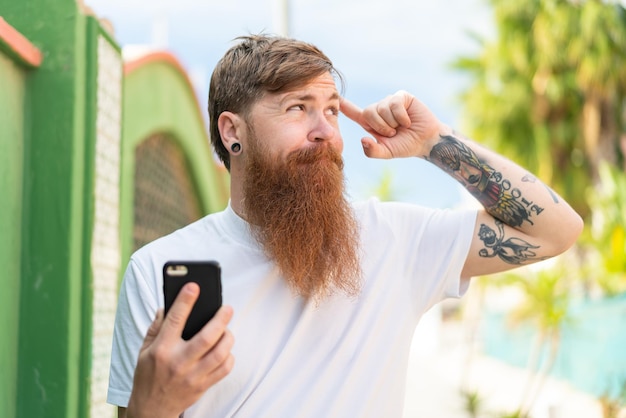 Hombre pelirrojo con barba usando teléfono móvil al aire libre con dudas y con expresión facial confusa