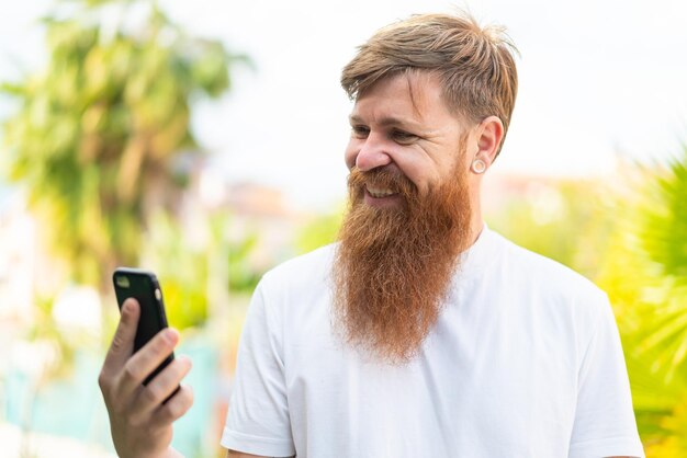 Hombre pelirrojo con barba usando teléfono móvil al aire libre con expresión feliz