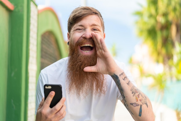 Hombre pelirrojo con barba usando teléfono móvil al aire libre gritando con la boca abierta