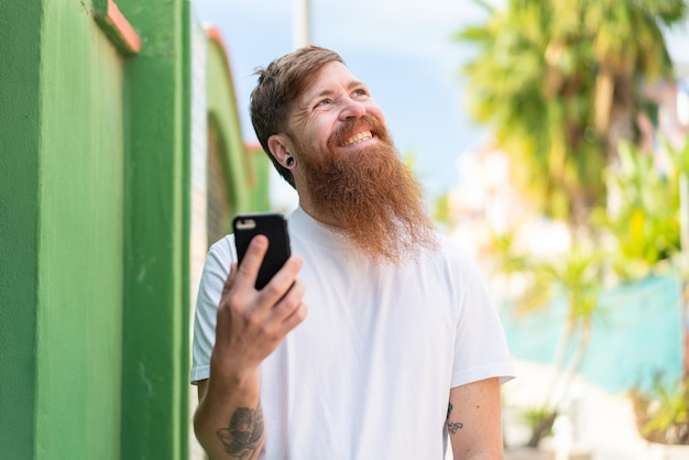 Hombre pelirrojo con barba usando teléfono móvil al aire libre mirando hacia arriba mientras sonríe