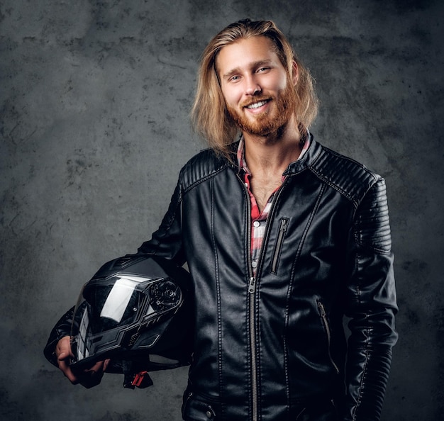 Hombre pelirrojo barbudo positivo con chaqueta de cuero sostiene casco de motocicleta sobre fondo gris.