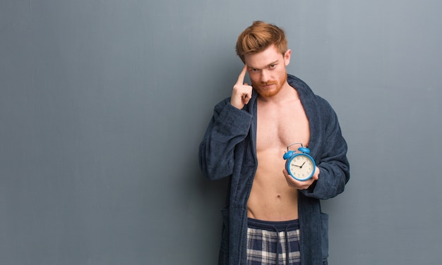 Hombre pelirrojo joven con pijama pensando en una idea. Él está sosteniendo un reloj despertador.