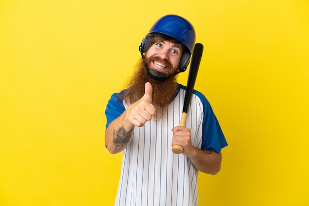 Hombre pelirrojo jugador de béisbol con casco y bate aislado sobre fondo amarillo dando un pulgar hacia arriba gesto