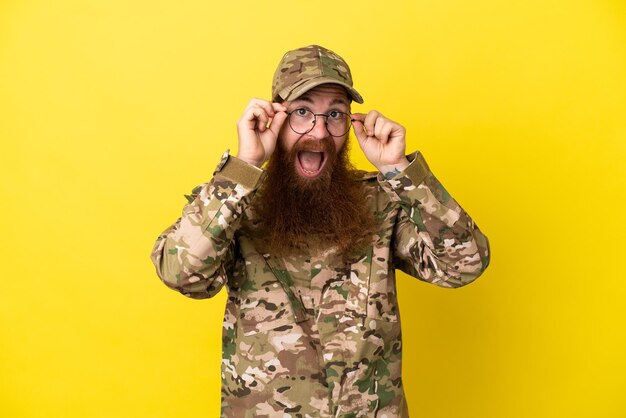 Hombre pelirrojo militar sobre aislado sobre fondo amarillo con gafas y sorprendido