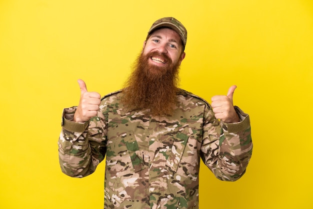 Hombre pelirrojo militar sobre aislado sobre fondo amarillo con gesto de pulgar hacia arriba y sonriente