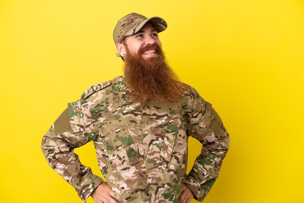Foto hombre pelirrojo militar sobre aislado sobre fondo amarillo posando con los brazos en la cadera y sonriendo
