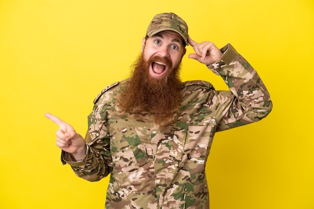 Foto hombre pelirrojo militar sobre aislado sobre fondo amarillo sorprendido y apuntando con el dedo hacia el lado