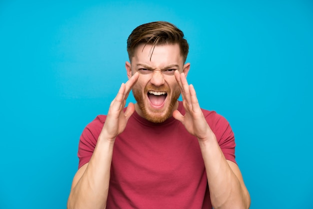 Hombre pelirrojo en pared azul aislada gritando con la boca abierta