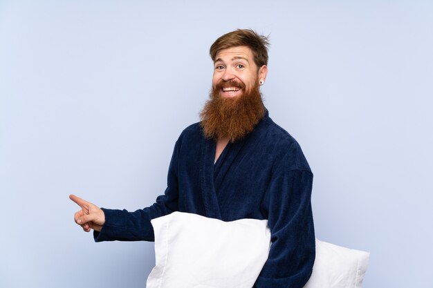 Hombre pelirrojo en pijama sobre pared aislada sorprendido y apuntando con el dedo a un lado