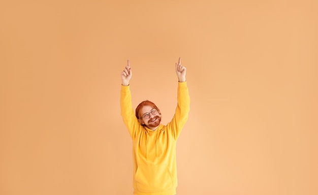 Un hombre pelirrojo sonriente con anteojos y una sudadera con capucha amarilla sobre un fondo amarillo levanta las manos hacia el espacio libre apuntándolo con las manos arriba Copiar espacio
