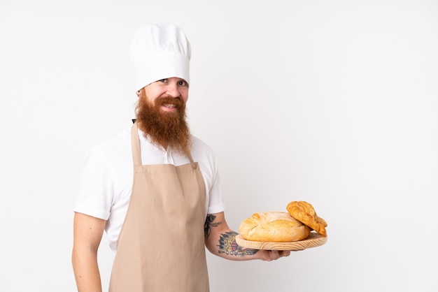 Hombre pelirrojo en uniforme de chef. Hombre panadero sosteniendo una mesa con varios panes sonriendo mucho