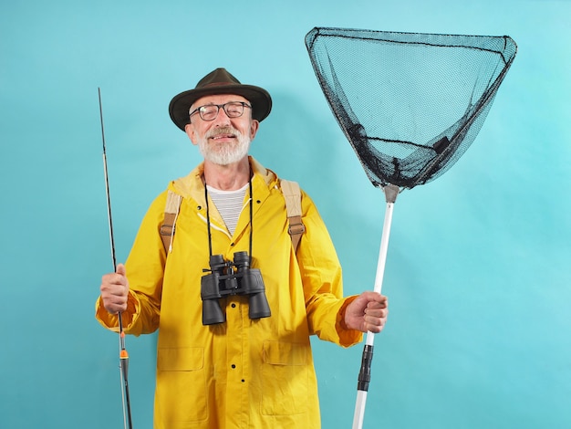 Hombre de pelo blanco con barba vestido con un impermeable amarillo tiene una caña de pescar y una red sobre un fondo aislado