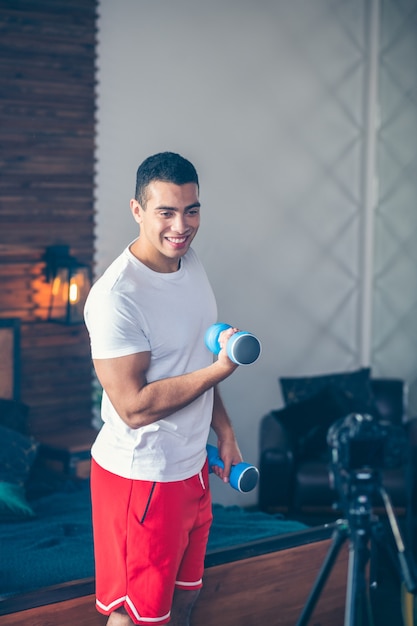 Hombre de pelo oscuro fuerte en pantalones cortos rojos sonriendo muy bien trabajando con mancuernas