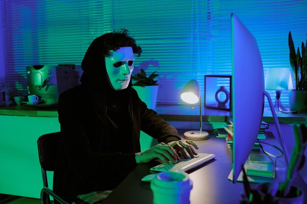 Hombre de pelo rizado con máscara blanca escondiendo su cara mientras programa virus a través de computadora en una habitación iluminada por
