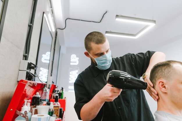 Hombre peluquero con una máscara médica y con un secador de pelo en las manos hace un corte de pelo para un cliente en una barbería Trabajo de barbería durante la cuarentena