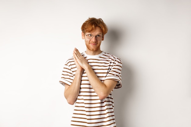 Foto hombre pensativo con cabello rojo frotándose las manos y mirando a la esquina superior derecha, teniendo una idea, de pie sobre fondo blanco.