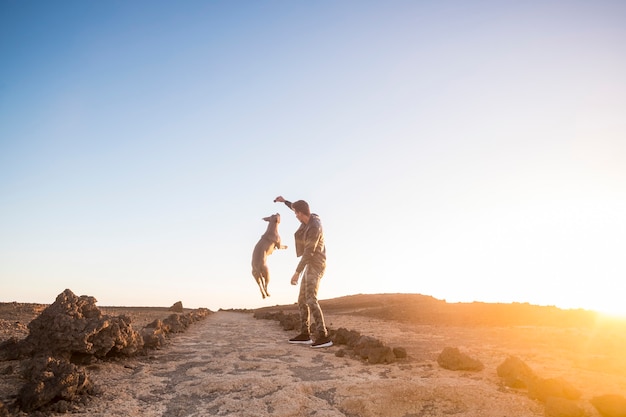 Hombre y perro juegan juntos saltando y divirtiéndose en actividades de ocio al aire libre durante una puesta de sol de color